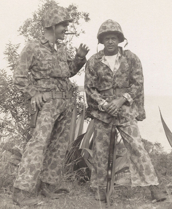 Two Marines in camouflage uniforms standing outdoors with trees in the background